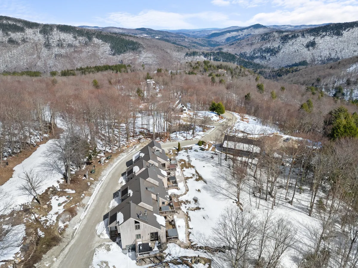 An exterior shot showing a row of condos nestled against a dramatic mountain range, with a winding road and snow-covered ground in view. The setting evokes the natural beauty of Killington and hints at easy access to hiking and biking trails as well as proximity to the mountains.