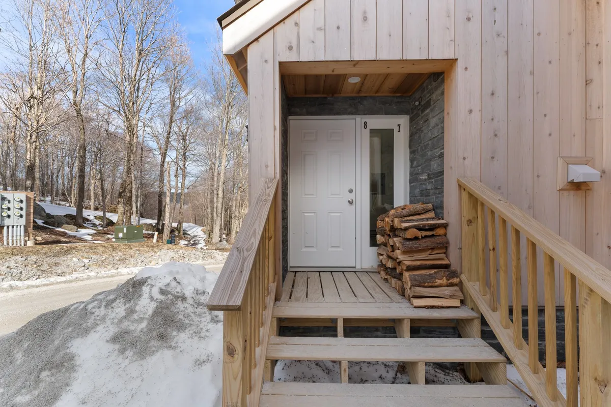 The image highlights the home's front door, stacked firewood, and wooden steps leading up from the snow. The overall mood is welcoming, suggesting easy access to outdoor spaces for activities like dog walks or playtime in the snow.