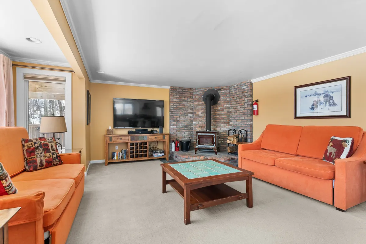 This image showcases the heart of the living area with inviting orange sofas surrounding a wood-burning stove and a television, all anchored by a cozy coffee table. The warm lighting and arrangement encourage gatherings for relaxation, conversation, or entertainment after a day on the slopes.