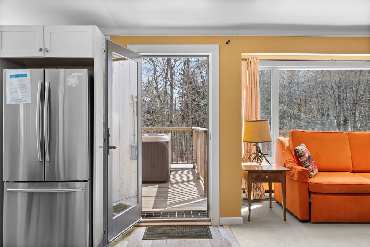 An interior shot featuring a cozy orange sofa, dining table, and a clear view of the private deck through large windows. The space is filled with natural light, suggesting a comfortable place to relax with your pet after time outdoors—and easy access to the deck for fresh air or quick clean-ups.