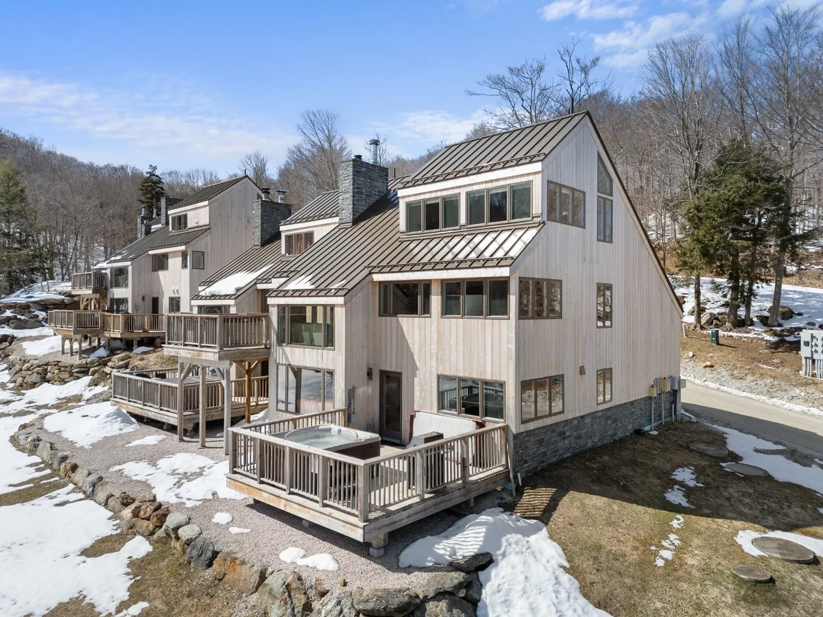 A modern wooden home with large windows and a balcony hot tub overlooks a snow-covered landscape. The inviting outdoor hot tub suggests relaxation after a day on the slopes, while the architecture and surrounding scenery highlight the cozy, upscale mountain experience.