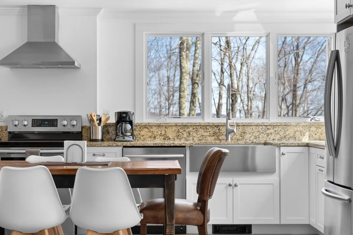 An open-concept kitchen and dining area features a granite countertop, stainless steel appliances, a wooden dining table set for a meal, and large windows letting in ample sunlight. The design highlights both cooking and shared dining experiences.