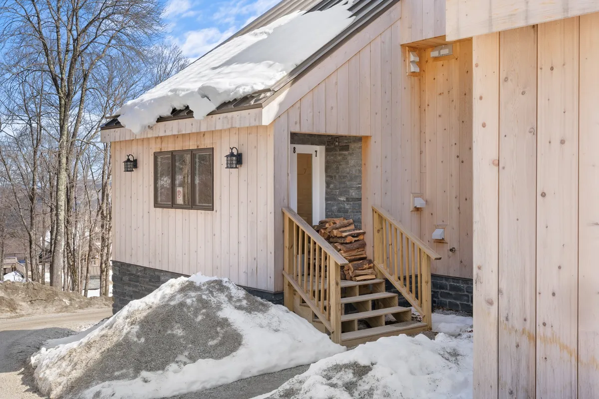 This exterior image captures a snow-covered roof, a welcoming wooden entrance, and a neatly stacked pile of firewood. The winter landscape and inviting entrance evoke a sense of adventure and warmth, hinting at a cozy retreat after outdoor activities.