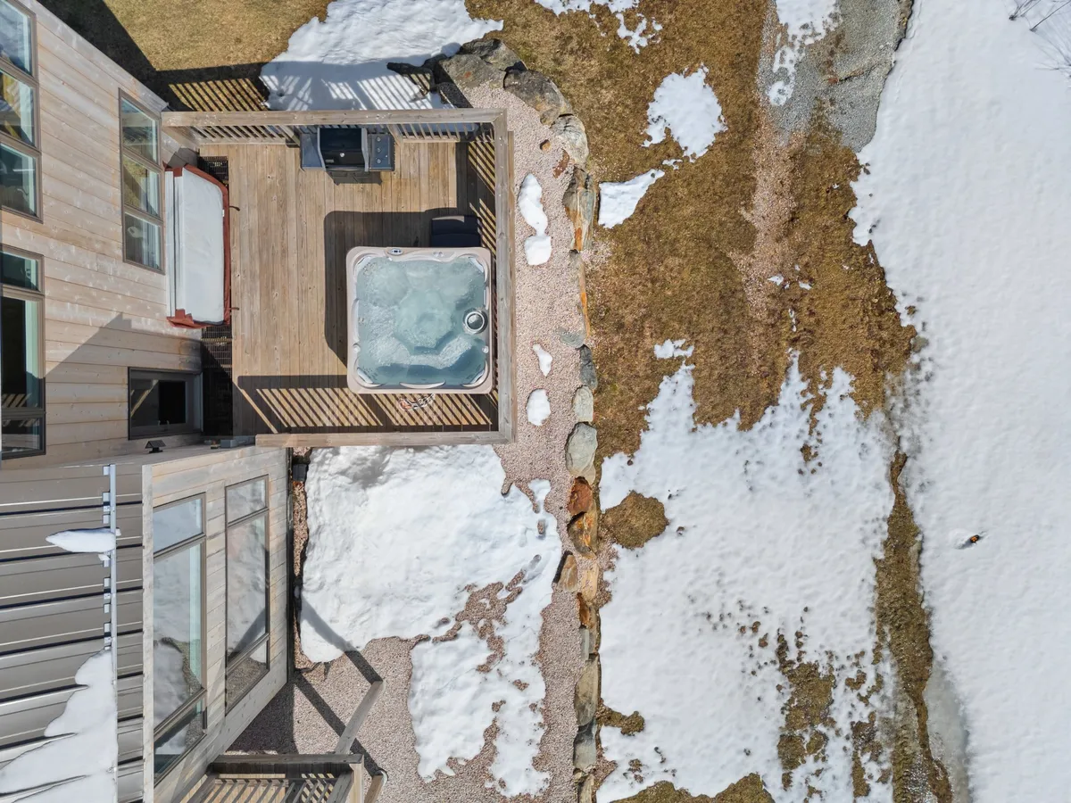 This exterior image features a private deck with a hot tub, surrounded by a mix of snow and grass, indicating it is usable across multiple seasons. The deck invites gatherings, relaxation, and al fresco dining, while the open natural setting hints at immediate access to outdoor activities.