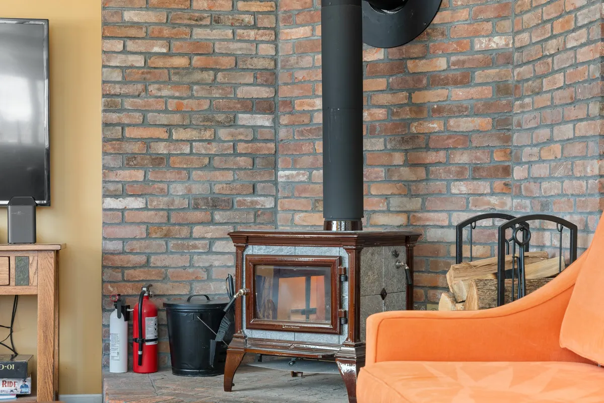 A welcoming corner of the living area featuring a wood stove, bright orange sofa, and a TV set against a brick wall. The décor and arrangement invite guests to snuggle up with blankets, play games, or have heartfelt conversations by the fire, all within a cheerful, pet-friendly setting.