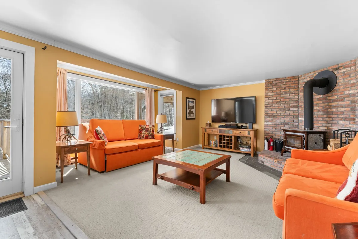 The image showcases a warm and inviting living room with vibrant orange sofas arranged around a classic brick fireplace. Sunlight streams through large windows, creating a bright, welcoming atmosphere ideal for relaxing after a day on the slopes.