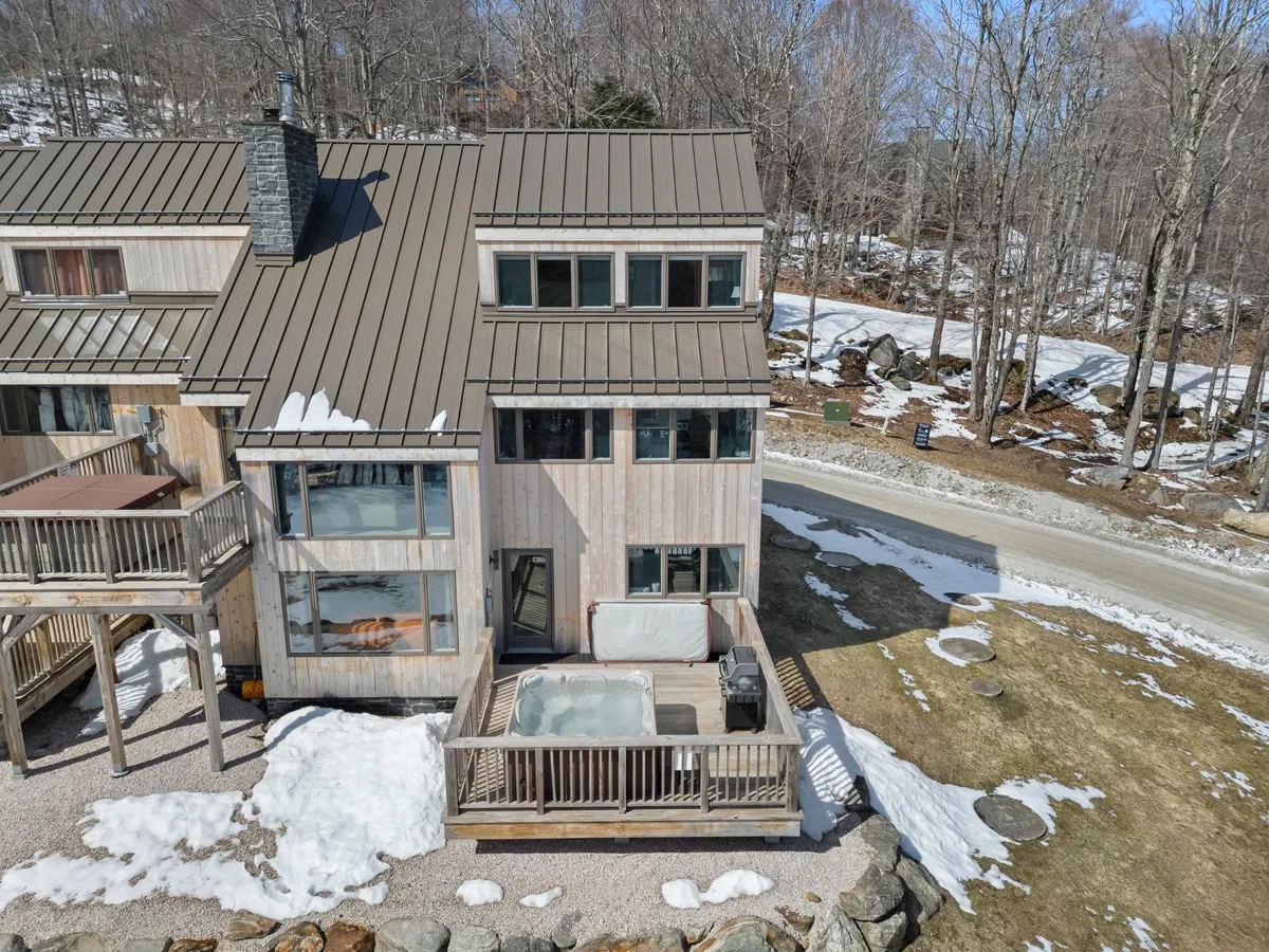 A cozy exterior view of the deck with a hot tub, wooden siding, snow on the ground, and large windows. The hot tub is the focal point, inviting guests to relax under the stars while surrounded by natural beauty.