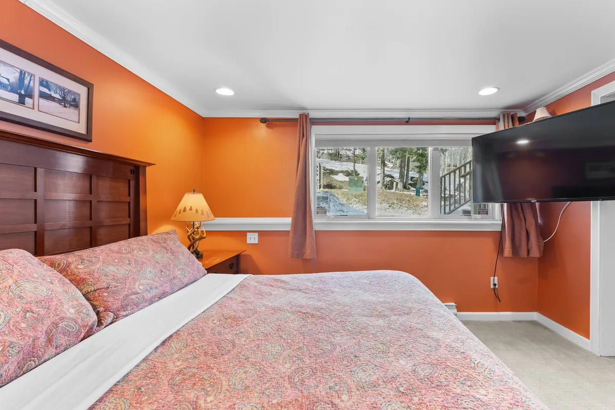 This image features a bed with patterned bedding, a wooden headboard, a decorative lamp, and a large window with a scenic view. The setting is inviting and comfortable, creating a restful ambiance ideal for relaxation after a busy day.