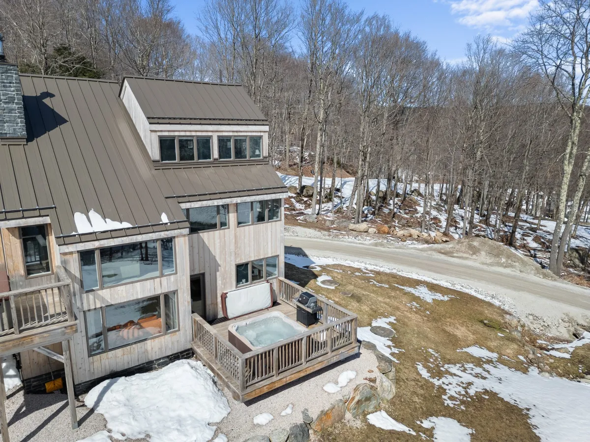 This image displays a private wooden deck nestled among trees, featuring a hot tub as its main focal point. The scene feels serene and secluded, with inviting large windows suggesting a direct connection to the cozy indoor living space. Fresh snow blankets the landscape, hinting at wintertime relaxation, while the hot tub appears ready for a romantic soak under the stars.